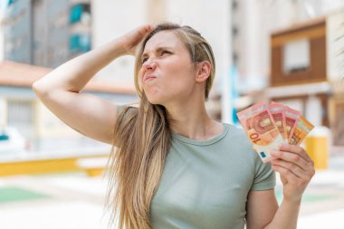 Genç sarışın kadın açık havada çok para alıyor. Şüpheleri var ve kafası karışık.