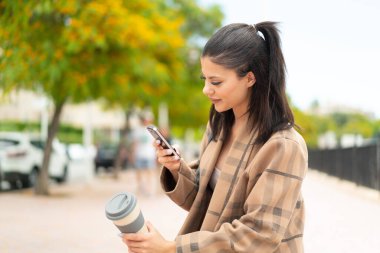 Dışarıda cep telefonu kullanan ve elinde kahve tutan genç ve güzel bir kadın.