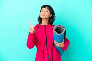 Mavi arka planda bir minder tutarken yoga derslerine giden genç kız gülümsüyor ve zafer işareti gösteriyor.