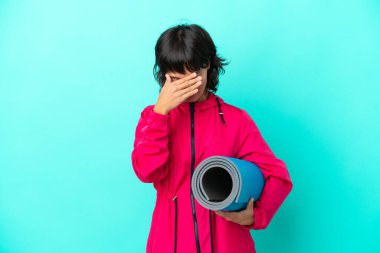 Mavi arka planda, yorgun ve hasta bir ifadeyle izole edilmiş bir minderi tutarak yoga derslerine giden genç kız.