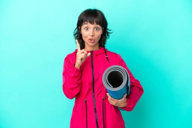 Mavi arka planda bir minder tutarak yoga derslerine giden genç kız çözümü bir parmağını kaldırarak bulmayı umuyor.