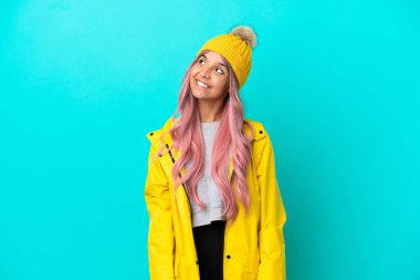 Pembe saçlı, yağmura dayanıklı bir ceket giyen genç bir kadın mavi arka planda izole edilmiş bir şekilde yukarı bakarken bir fikir düşünüyor.