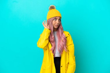 Pembe saçlı, yağmura dayanıklı bir ceket giyen, mavi arka planda izole edilmiş genç bir kadın kulağına elini sürerek bir şeyler dinliyor.