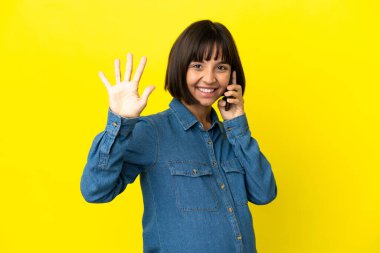 Sarı arka planda cep telefonu kullanan hamile kadın parmaklarıyla beşi sayıyor.