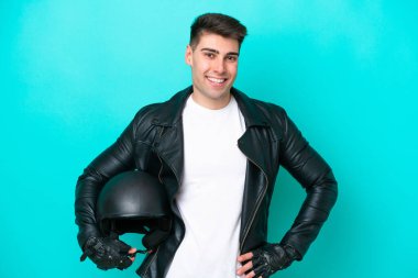 Motosiklet kasklı genç beyaz adam mavi arka planda izole edilmiş. Kolları kalçasında ve gülümsüyor.