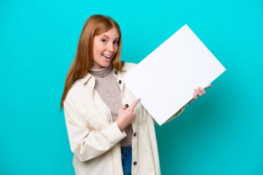 Genç kızıl saçlı kadın mavi arka planda izole edilmiş. Elinde mutlu bir ifadeyle boş bir levha var.