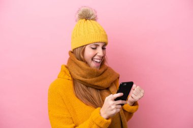 Kışlık ceket giyen genç kızıl saçlı kadın. Pembe arka planda izole edilmiş ve telefon zafer pozisyonunda.