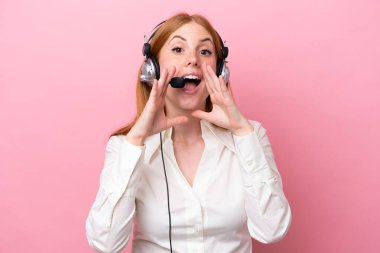 Tele pazarlamacı kızıl saçlı kadın pembe arka planda izole edilmiş bir kulaklıkla çalışıyor bağırıyor ve bir şey duyuruyor.