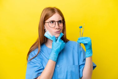 Genç kızıl saçlı dişçi kadın sarı arka planda izole edilmiş. Şüpheleri var.