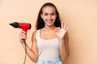 Elinde bej arka planda izole edilmiş saç kurutma makinesi tutan genç bir kadın parmaklarıyla tamam işareti gösteriyor.