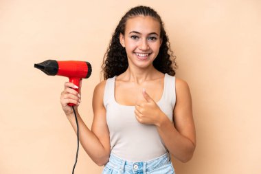 Elinde bej arka planda izole edilmiş saç kurutma makinesi tutan genç bir kadın. Başparmakları havada çünkü iyi bir şey oldu.