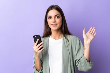 Mor arka planda izole edilmiş cep telefonu kullanan beyaz bir kadın mutlu bir ifadeyle selamlıyor.