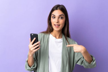 Mor arka planda izole edilmiş cep telefonu kullanan beyaz bir kadın. Şaşırtıcı bir yüz ifadesi var.