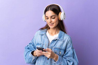 Genç beyaz kadın mor arka planda izole edilmiş müzik dinliyor ve cep telefonu arıyor.