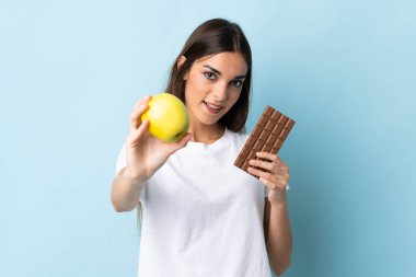 Kafkasyalı genç bir kadın mavi arka planda izole edilmiş. Bir elinde çikolata tableti, diğer elinde elma var.