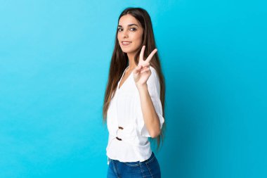 Mavi arka planda izole edilmiş genç beyaz kadın gülümsüyor ve zafer işareti gösteriyor.