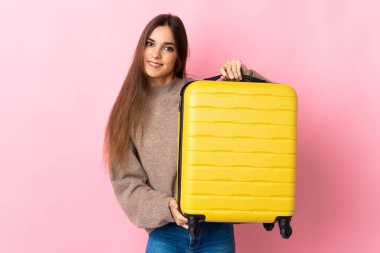 Pembe arka planda izole edilmiş genç beyaz kadın seyahat valiziyle geziyor.