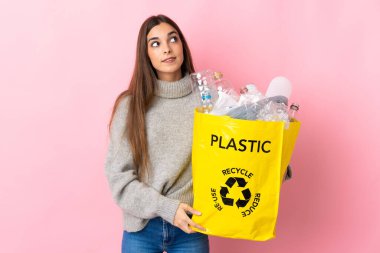 Pembe arka planda izole edilmiş bir şekilde plastik şişelerle dolu bir çanta taşıyan genç beyaz kadın bir fikir ararken...