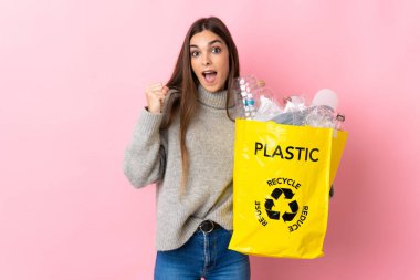 Pembe arka planda izole edilmiş plastik şişelerle dolu bir çanta taşıyan beyaz bir kadın zaferi kutluyor.