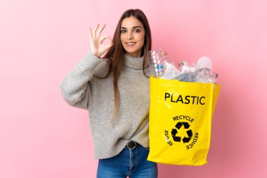 Pembe arka planda izole edilmiş geri dönüşüm için elinde plastik şişelerle dolu bir çanta tutan beyaz bir kadın parmaklarıyla tamam işareti gösteriyor.