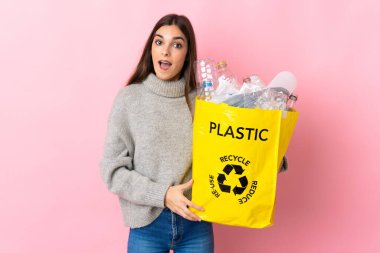 Elinde plastik şişelerle dolu bir çanta tutan beyaz bir kadın. Pembe arka planda izole edilmiş, şaşırmış bir yüz ifadesiyle geri dönüşüm için.