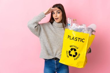 Elinde plastik şişelerle dolu bir çanta tutan beyaz bir kadın. Pembe arka planda izole edilmiş, sürpriz bir ifadeyle geri dönüşüm için.