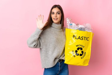 Pembe arka planda izole edilmiş plastik şişelerle dolu bir çanta taşıyan beyaz bir kadın beş parmağıyla sayıyor.