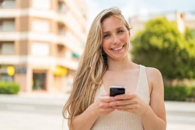 Dışarıdaki genç sarışın kadın cep telefonuyla mesaj yolluyor.