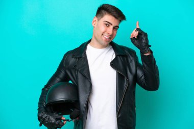 Motosiklet kasklı genç beyaz adam mavi arka planda izole edilmiş ve en iyinin işareti olarak parmağını kaldırıyor.