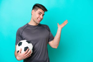 Kafkasyalı genç futbolcu, mavi arka planda izole edilmiş. Davet etmek için ellerini yana uzatıyor.