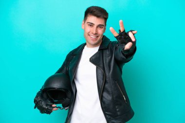 Motosiklet kasklı genç beyaz adam mavi arka planda izole edilmiş gülümsüyor ve zafer işareti gösteriyor.