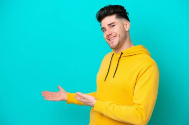 Mavi arka planda izole edilmiş genç beyaz adam, davet edilmek için ellerini yana uzatıyor.