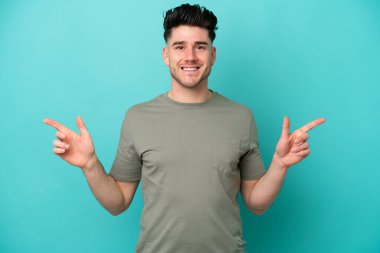 Mavi arka planda izole edilmiş genç beyaz adam parmak uçlarını işaret ediyor ve mutlu.