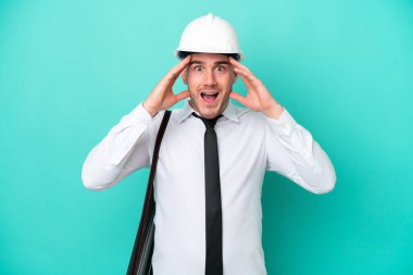 Miğferli ve elinde mavi arkaplanda sürpriz bir ifadeyle izole edilmiş planları olan genç bir beyaz adam.