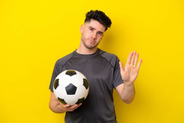 Yakışıklı genç futbolcu sarı arka planda izole edilmiş. Durma hareketi yapıyor ve hayal kırıklığına uğruyor.