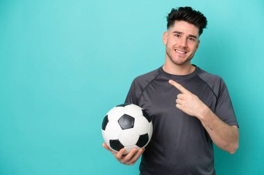 Kafkasyalı genç adam mavi arka planda, futbol topuyla izole edilmiş ve yan tarafı işaret ediyor.