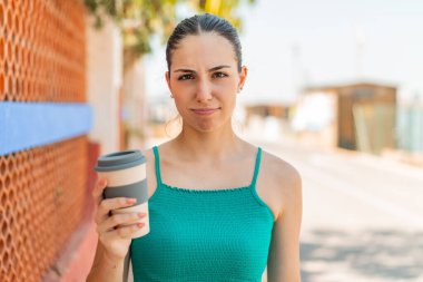 Genç ve güzel bir kadın üzgün bir ifadeyle açık havada kahve tutuyor.