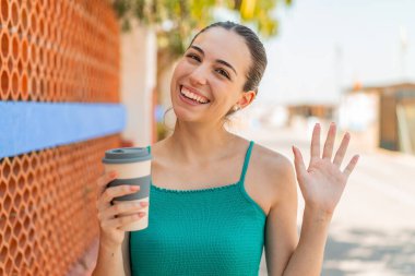 Genç ve güzel bir kadın açık havada kahvesini yudumlarken mutlu bir ifadeyle selamlıyor.