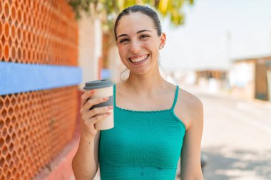 Genç ve güzel bir kadın elinde kahveyle dışarı çıkıp gülümsüyor.