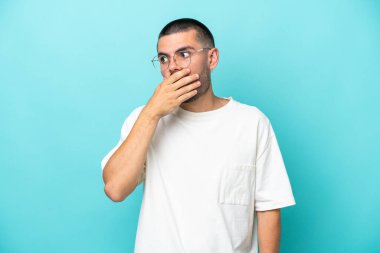 Mavi arka planda izole edilmiş genç beyaz adam yan tarafa bakarken sürpriz jest yapıyor.