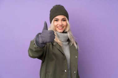 Kış şapkası takmış genç sarışın kız. Mor arka planda. Başparmakları havada.