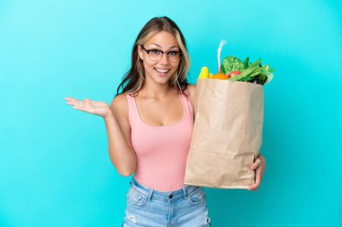 Young Russian woman holding a grocery shopping bag isolated on blue background with shocked facial expression