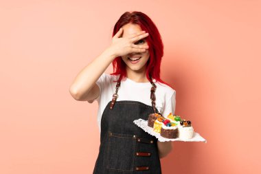 Pasta şefi pembe arka planda izole edilmiş bir kek tutuyor gözleri elle kaplıyor ve gülümsüyor.