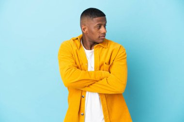 Mavi arka planda izole edilmiş genç Latin adam kollarını kavuşturuyor.