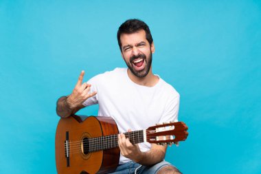 İzole edilmiş mavi arka planda gitar çalan genç bir adam kaya jesti yapıyor.
