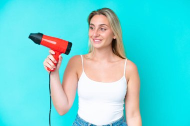 Kafkasyalı genç bir kadın elinde saç kurutma makinesiyle mavi arka planda mutlu bir ifadeyle izole edilmiş.