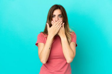 Mavi arka planda izole edilmiş genç beyaz kadın elleriyle ağzını kapatıyor.