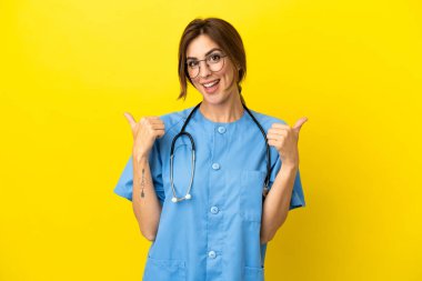 Doktor kadın, sarı arka planda izole edilmiş. Baş parmakları havada ve gülümsüyor.