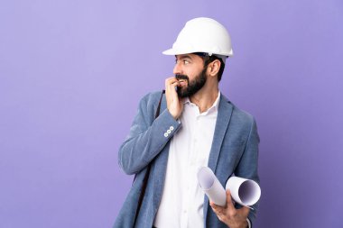 Miğferli genç bir mimar, izole edilmiş mor arka plan üzerinde planları var. Gergin ve eli ağıza vermeye korkuyor.
