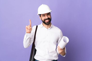 Miğferli genç bir mimar. Mor arka planda izole edilmiş planlar tutuyor. En iyinin işareti olarak parmağını kaldırıyor.